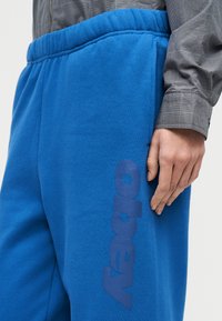 Άτομο που φορά φωτεινό μπλε φόρμα με το λογότυπο "obey" στον μηρό και γκρι καρό μακρυμάνικο πουκάμισο, με το χέρι ακουμπισμένο στο πλάι.