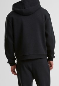 Uomo che indossa una felpa nera semplice e pantaloni neri abbinati, visto di spalle su uno sfondo neutro.