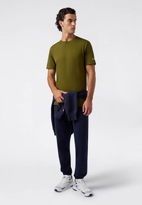 Olive green short-sleeve t-shirt with logo on sleeve, paired with navy sweatpants. Model holds a navy sweater. White sneakers complete the look.
