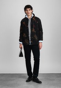 Giacca in velluto a coste nera con motivi floreali colorati, indossata sopra un maglione grigio a maglia, abbinata a pantaloni scuri e scarpe nere, mentre tiene una borsa a mano nera.