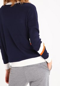 Mörkblå stickad tröja med vita och orangea randiga detaljer på ärmarna. Ribbad mudd och fåll. Mjuk textur, avslappnad passform.