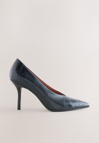 Chaussure noire en cuir à talon haut, bout pointu, motif carré cousu et talon stiletto sur fond neutre.