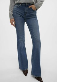 Jean évasé bleu clair en denim, comportant cinq poches et des coutures discrètes, associé à des escarpins marron pointus.