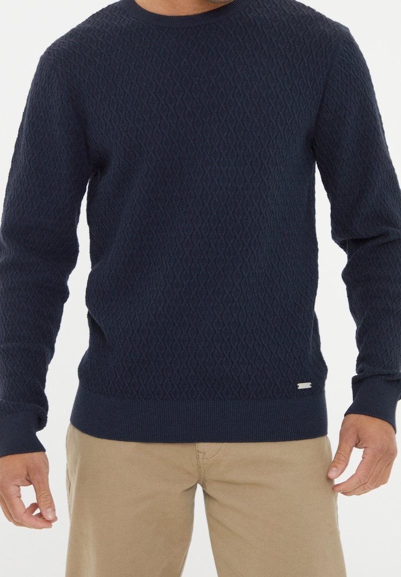 Homme portant un pull à col rond texturé bleu marine et un pantalon beige, debout les bras détendus le long du corps sur un fond blanc.