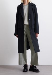 Abrigo largo de lana negra con solapa, dos bolsillos frontales y botones. Lleva puesto un suéter gris claro y pantalones verde oliva. Zapatos negros.