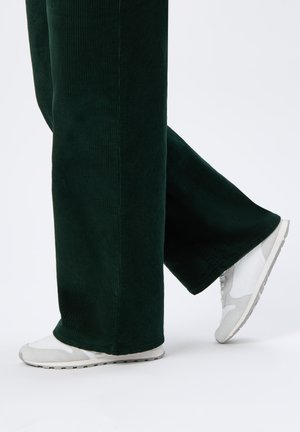 Leg-length dark green corduroy pants with white and gray sneakers, one foot flat, the other raised at the heel against a white background.