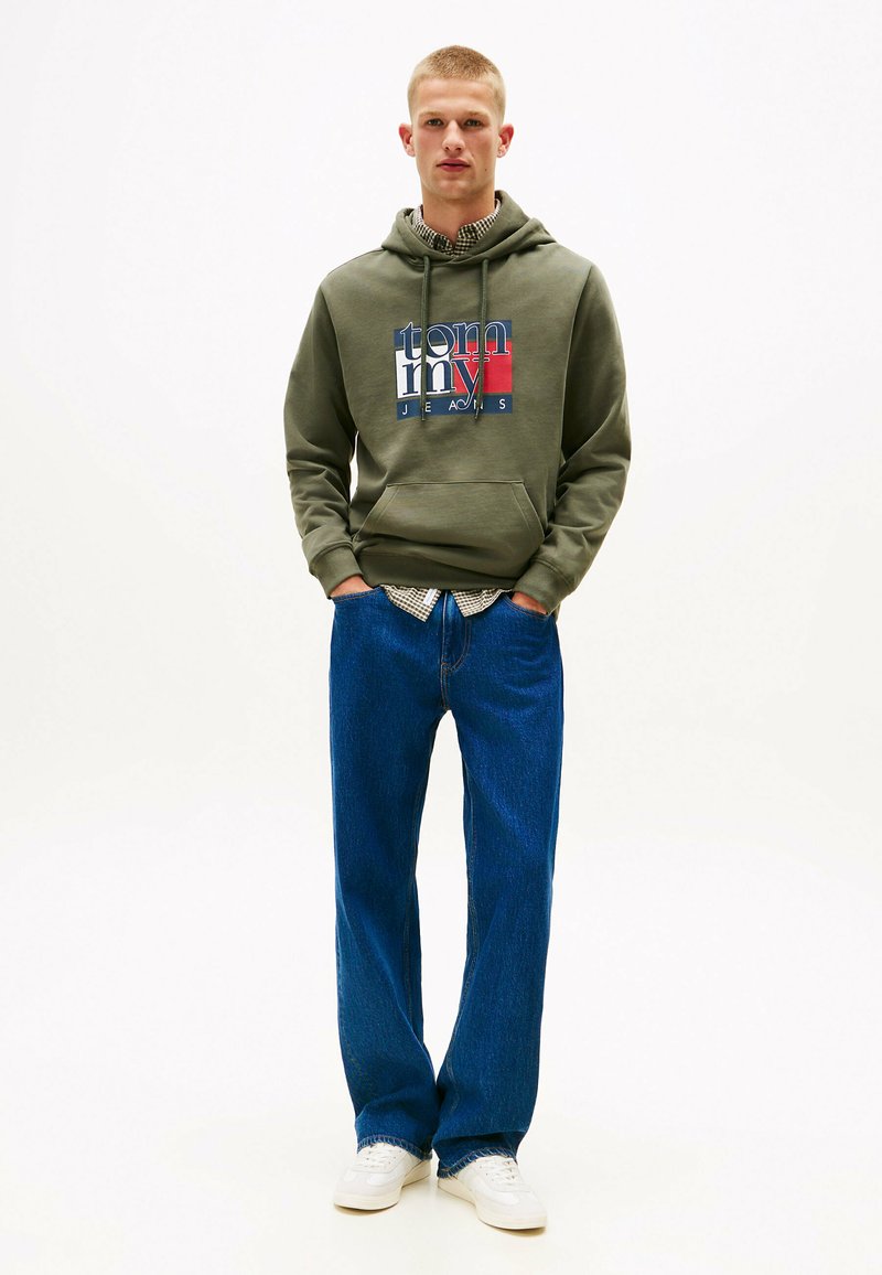 Hættetrøje i olivengrøn med logo-print, kombineret med løse blå denimjeans og hvide sneaks. Struktureret stof og afslappet pasform.