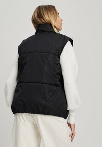 Juodas pūkinis liemenė be rankovių ir su aukštu apykakle; pasižymi siuvinėjimu ir lygiu paviršiumi, derinama su balta raštuota megztine.