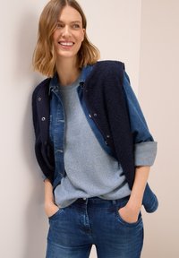 Eine lächelnde Frau lehnt an einer Wand und trägt ein mehrlagiges blaues Outfit mit einem Jeanshemd, einem Strickpullover, einer Fleeceweste und Jeans, die Hände in den Taschen.