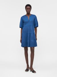 Femme portant une robe bleue à volants avec des manches bouffantes jusqu'au coude et des sandales noires à talons bas et pieds ouverts, debout sur un fond blanc.