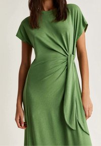 Femme portant une robe verte à manches courtes avec un détail de nœud sur le côté, montrant son ventre et ses bras devant un fond uni.