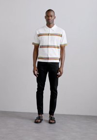 Valkoinen lyhythihainen paita, jossa on diamond-kuvio ja moniväriset raidat, yhdistetty mustiin slim fit -housuihin ja tummiin sandaaleihin.