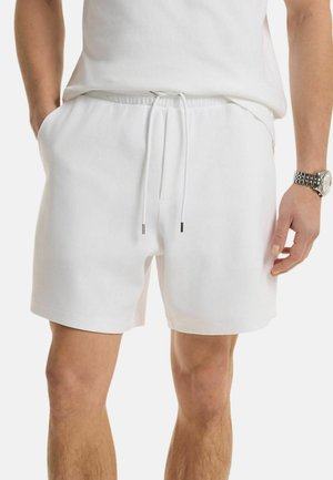 Uomo che indossa pantaloncini bianchi semplici con coulisse e tasche, abbinati a una camicia bianca e un orologio da polso argentato al polso destro.