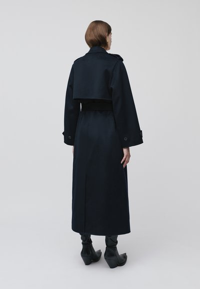 Długi granatowy płaszcz z paskiem w talii, wyraźnie zarysowanymi ramionami i szerokimi rękawami. Gładka tkanina, rozporek po bokach oraz guziki na mankietach.