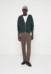 Hoodie zippé vert foncé, t-shirt blanc, pantalon marron et chaussures noires. Le hoodie présente un petit logo ; la casquette est beige clair.