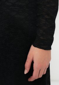 Μαύρο φόρεμα με υφή λουλουδιών, με εφαρμοστή σχεδίαση και μακρυά, διάφανα μανίκια. Κοντινό πλάνο ενός χεριού που στηρίζεται στο ύφασμα.