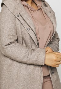 Světlohnědý kabát z texturované tkaniny, s předním zipem, knoflíkovými detaily a kapucí, vrstvený přes odpovídající růžové mikině.