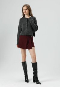 Cardigan en maille gris avec boutons, associé à une mini-jupe bordeaux avec un pli et un détail en chaîne. Bottes noires montantes à talons carrés.