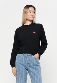 Black long-sleeve t-shirt with a small red heart logo on the chest, paired with blue vertically striped jeans. Plain background.