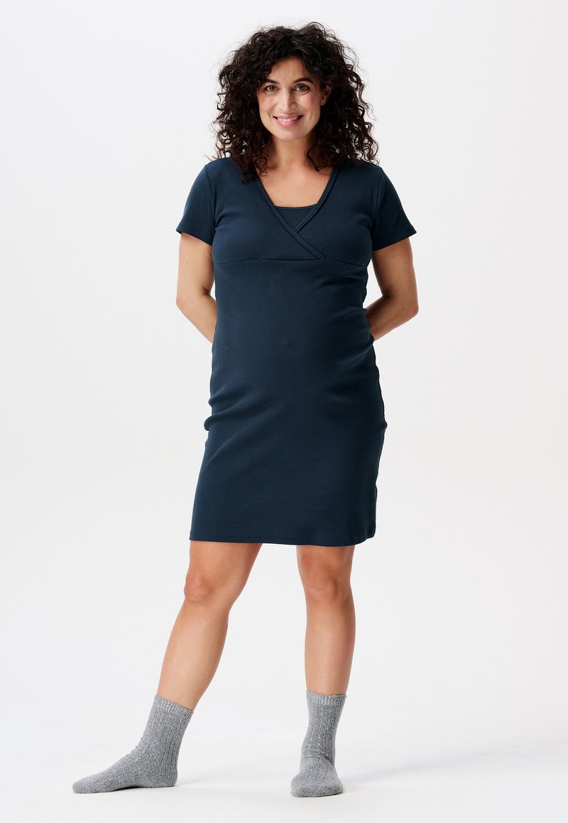 Granatowa sukienka ciążowa z krótkimi rękawami i krzyżującym się wzorem przy dekolcie, w połączeniu z jasnoszarymi, wełnianymi skarpetami. Gładki materiał.