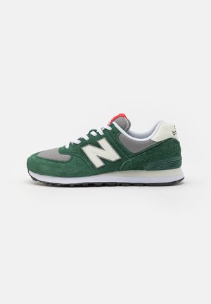Grüne Wildleder- und graue Mesh-Sneaker mit weißen Akzenten, ausgestattet mit einem markanten "N"-Logo und einem roten Fersenbereich. Klassisches Design der Gummisohle.