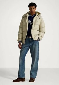 Mladic odjeven u bež kaput s kapuljačom, mornarsko plavi džemper s bijelim tekstom, plave traperice i smeđe cipele, stoji s rukama u džepovima.