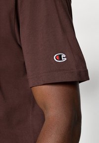 Brun bomulds t-shirt med korte ærmer og en buet nederkant, med et rødt og sort broderet logo på venstre ærme.
