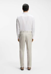 Mand stående med ryggen til, iført en hvid langærmet skjorte, beige bukser og brune sko på en ensfarvet hvid baggrund.