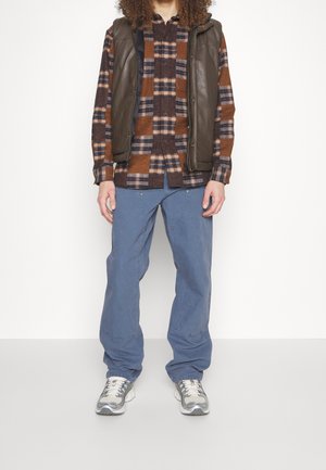 Personne portant une chemise à carreaux marron et bleu marine, une doudoune sans manches marron foncé, un pantalon bleu et des baskets blanches, debout devant un fond blanc.