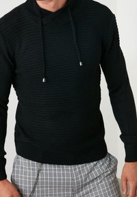 Pull noir en maille avec un col montant, arborant un design asymétrique et des cordons ajustables. Motif texturé avec des poignets côtelés.
