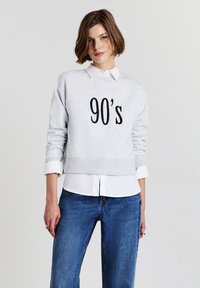 Felpa grigia con la scritta "90's" stampata in nero, caratterizzata da un collo rotondo e polsini a costine, sovrapposta a una camicia bianca con colletto. In evidenza jeans in denim.