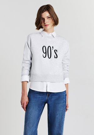 Grauer Sweatshirt mit dem Aufdruck "90's" in Schwarz, mit runder Halslinie und gerippten Bündchen, über einem weißen Hemd mit Kragen getragen. Präsentiert werden Jeans aus Denim.