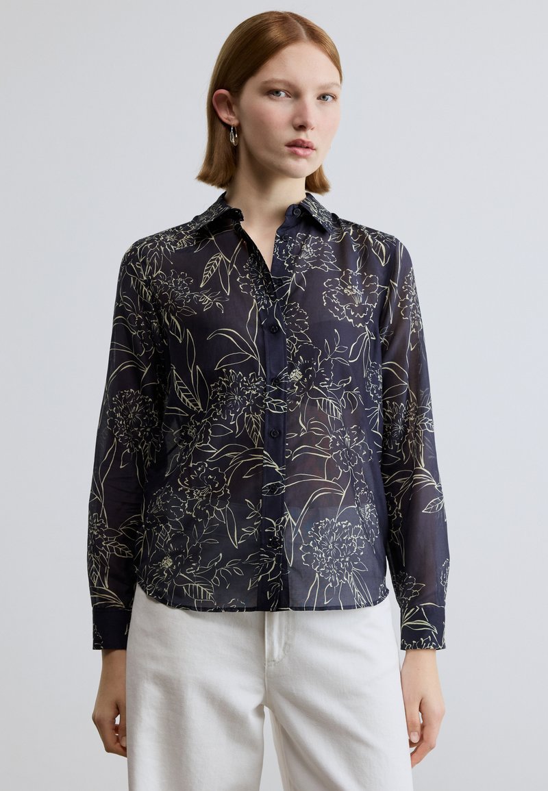 Camisa transparente azul-marinho com padrão de desenho floral em linhas, fecho de botões e colarinho clássico. Mangas compridas com detalhe em punho, combinada com calças brancas.