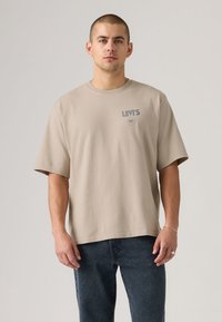 T-shirt bege Levi's 501, mangas curtas, gola redonda, tecido macio, logo frontal em cinza, combinada com jeans denim escuro.