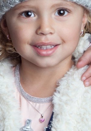 Kind met krullend blond haar, draagt een grijze pet, een witte fluffy jas en een roze top. Heeft een zilveren ketting met een hartvormige hanger.