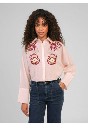 Femme aux cheveux bouclés portant un chemisier rose clair avec une broderie florale rouge et jaune, rentrée dans un jean bleu foncé taille haute.