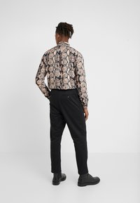 Dlouhý rukáv košile s béžovým a černým vzorem hada, doplněná černými kalhotami a černými botami. Hladká textura a přiléhavý střih.