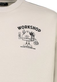 Light beige sweatshirt featuring a graphic print of a cartoon handyman with tools, labeled "WORKSHOP" and "AVAILABLE TO FIX EVERYTHING."