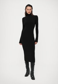 Črna oprijeta turtleneck obleka iz gladkega, elastičnega materiala, z dolgimi rokavi in midi dolžino, v kombinaciji z črnimi škornji do kolen.