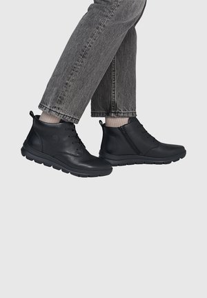 Person wearing black leather ankle boots with laces and side zippers, light beige socks, and gray denim jeans against a plain background.