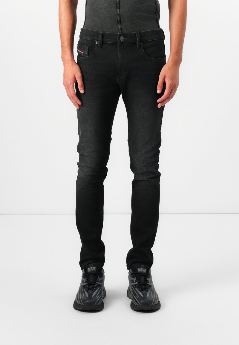 Jeans de mezclilla negra con un corte slim, que presentan un ligero desgaste, diseño clásico de cinco bolsillos y cierre de botón. Llevados con zapatillas atléticas negras.