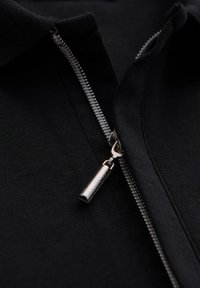 Černá košile na zip s vysokým límcem; obsahuje kovový zip a hladkou, strukturovanou tkaninu. Detailní záběr ukazuje zip a švy.