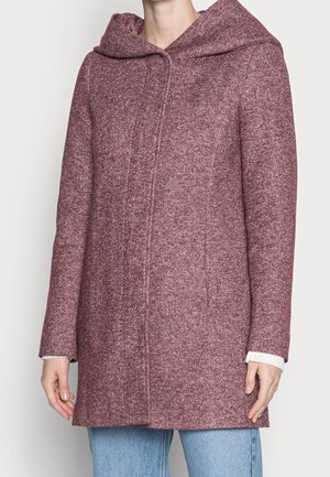 Femme portant un manteau texturé bordeaux avec une grande capuche par-dessus un pull blanc et un jean bleu clair, debout devant un fond uni.