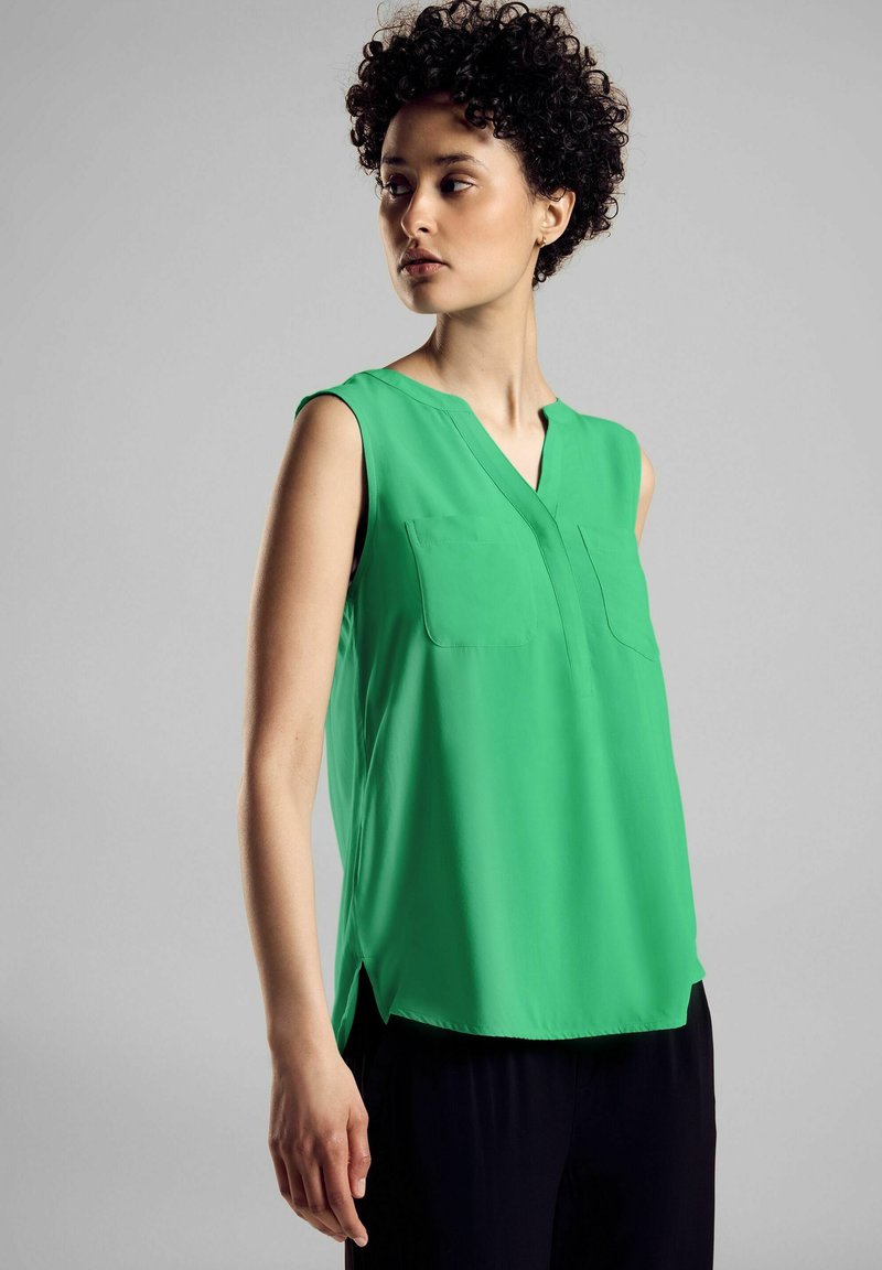 Vihreä ilman hihoja oleva bluus, jossa on V-pääntie, kaksi rintataskua ja pehmeä, sileä rakenne. Lievästi pussittava malli, jossa on sivuhalkiot.