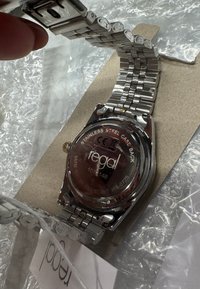 Silberne Uhr aus Edelstahl mit einem Metallarmband, rundem Gehäuse, transparentem Rücken und eingraviertem Logo auf rotem Hintergrund.