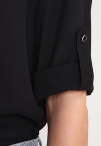 Maglietta nera con maniche arrotolate e dettaglio con bottone. Realizzata in materiale leggero, presenta una texture liscia e una vestibilità rilassata.