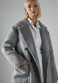 Grau melierter Wollmischmantel mit strukturierter Oberfläche, Reverskragen, Doppelknopfverschluss und seitlichen Taschen. Getragen über einem weißen Hemd.