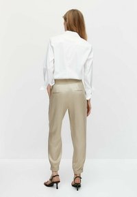 Kobieta odwrócona plecami, ubrana w białą bluzkę z długimi rękawami, beżowe satynowe spodnie na elastycznym pasku i czarne sandały na obcasie, na tle jednolitym.