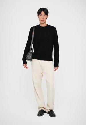 Black knit sweater, loose cream-colored pants, and black leather shoes. Model holds a black bag with a silver accent. Simple background.