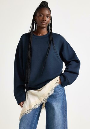 Pitkä punottu tukka, yllään ylisuuri tummansininen collegepaita, vaaleat farkut ja kermaisen värinen, pitsireunainen huivi vyötäröllä.